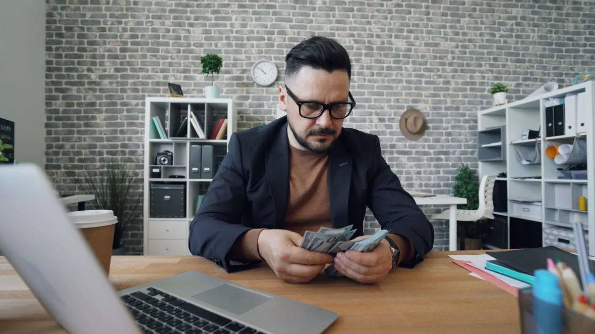 Mann im Sakko zählt am Schreibtisch vor einem Laptop Geldscheine