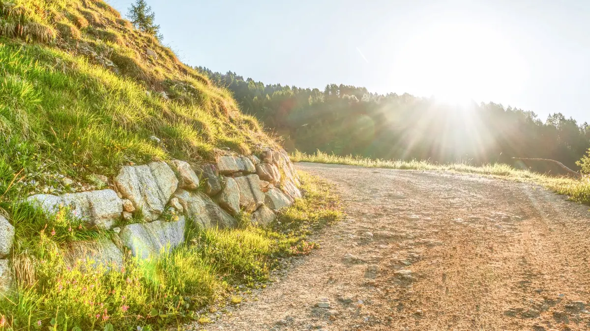 Sonnenaufgang über einem Feldweg mit Trockenmauer und Wiese am Hang
