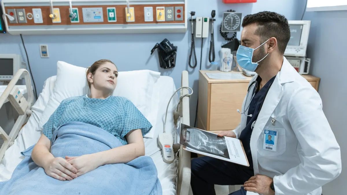 Arzt mit Mundschutz bespricht mit einer Patientin im Krankenbett die Behandlung