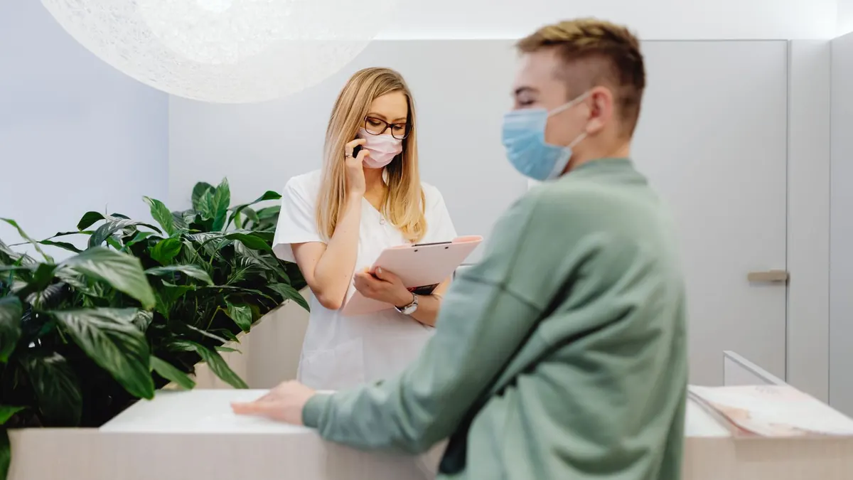 Patient mit Mundschutz steht am Empfangstresen einer Arztpraxis