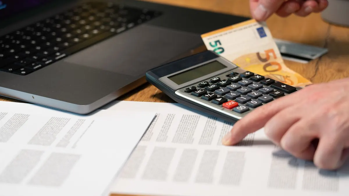 Hand zeigt auf Unterlagen neben Taschenrechner, Laptop und Fünfzig-Euro-Schein