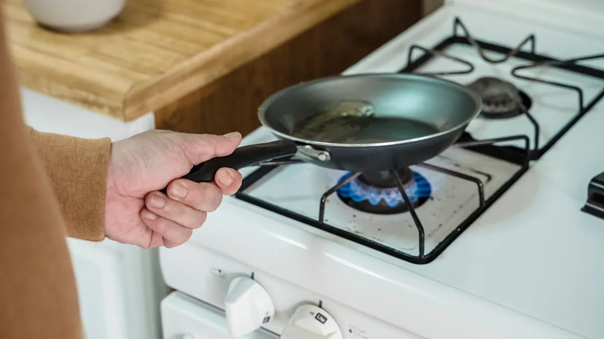 Hand hält eine Pfanne über einer blauen Gasflamme auf dem Herd
