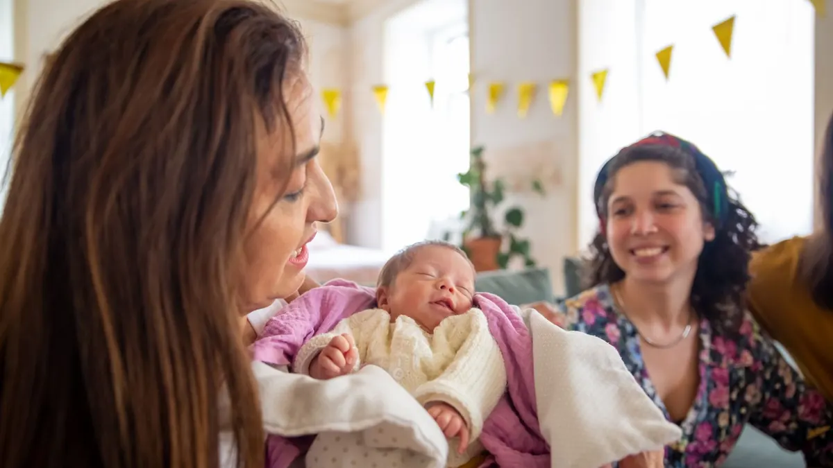 Mutter hält ihr Neugeborenes im Arm, daneben lächelt eine Frau