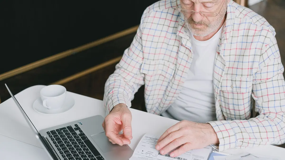 Älterer Mann prüft am Laptop Unterlagen mit einer Tasse Kaffee