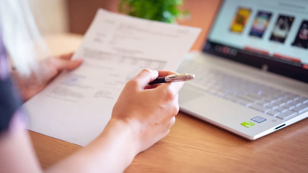 Hand hält einen Beleg mit Stift neben einem aufgeklappten Laptop
