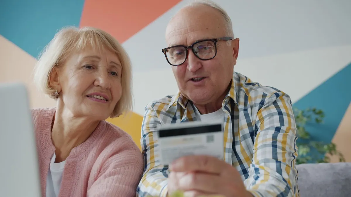 Älteres Paar schaut gemeinsam lächelnd auf eine Bankkarte im Wohnzimmer