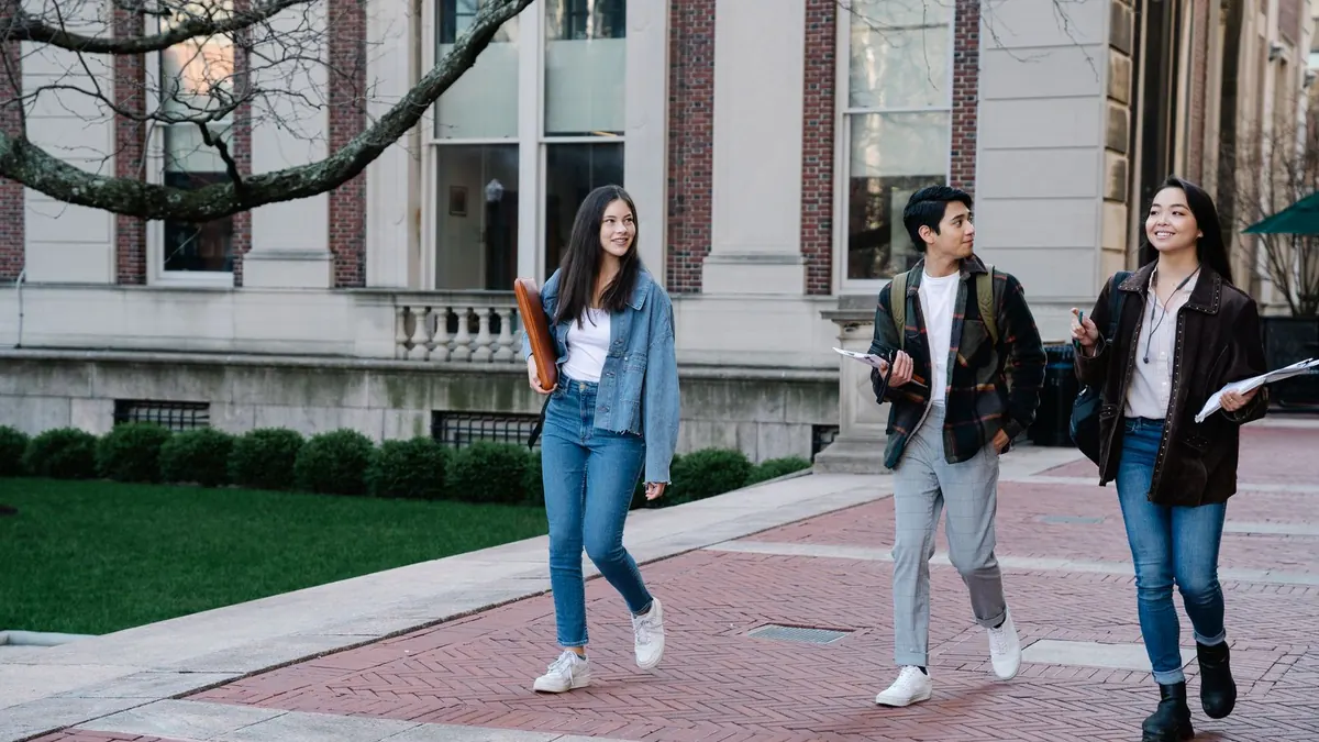 Drei Studierende gehen mit Taschen über einen Campus vor Universitätsgebäuden