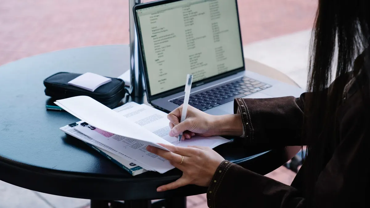 Frau macht am Tisch mit Laptop handschriftliche Notizen auf Unterlagen
